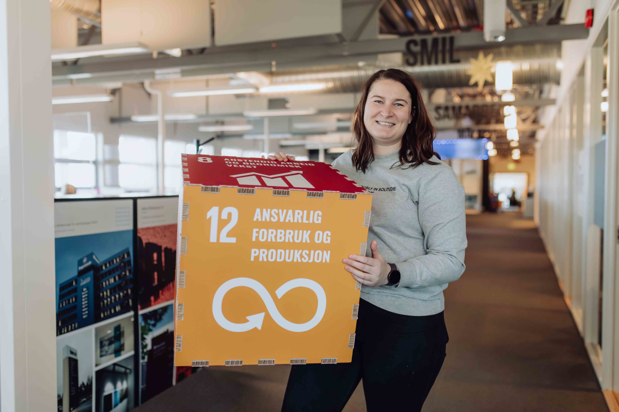 En ung dame holder en pappboks med ulike bærekraftsmål på. Mål nr. 12: ansvarlig forbruk og produksjon er vendt mot kamera.